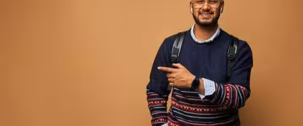 smiling happy indian student with backpack pointing his finger wall