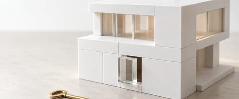 White architectural model of a modern house built from matte blocks, with one clear glass block pushed out of place to leave an opening, and a brass key lying unused on the table.